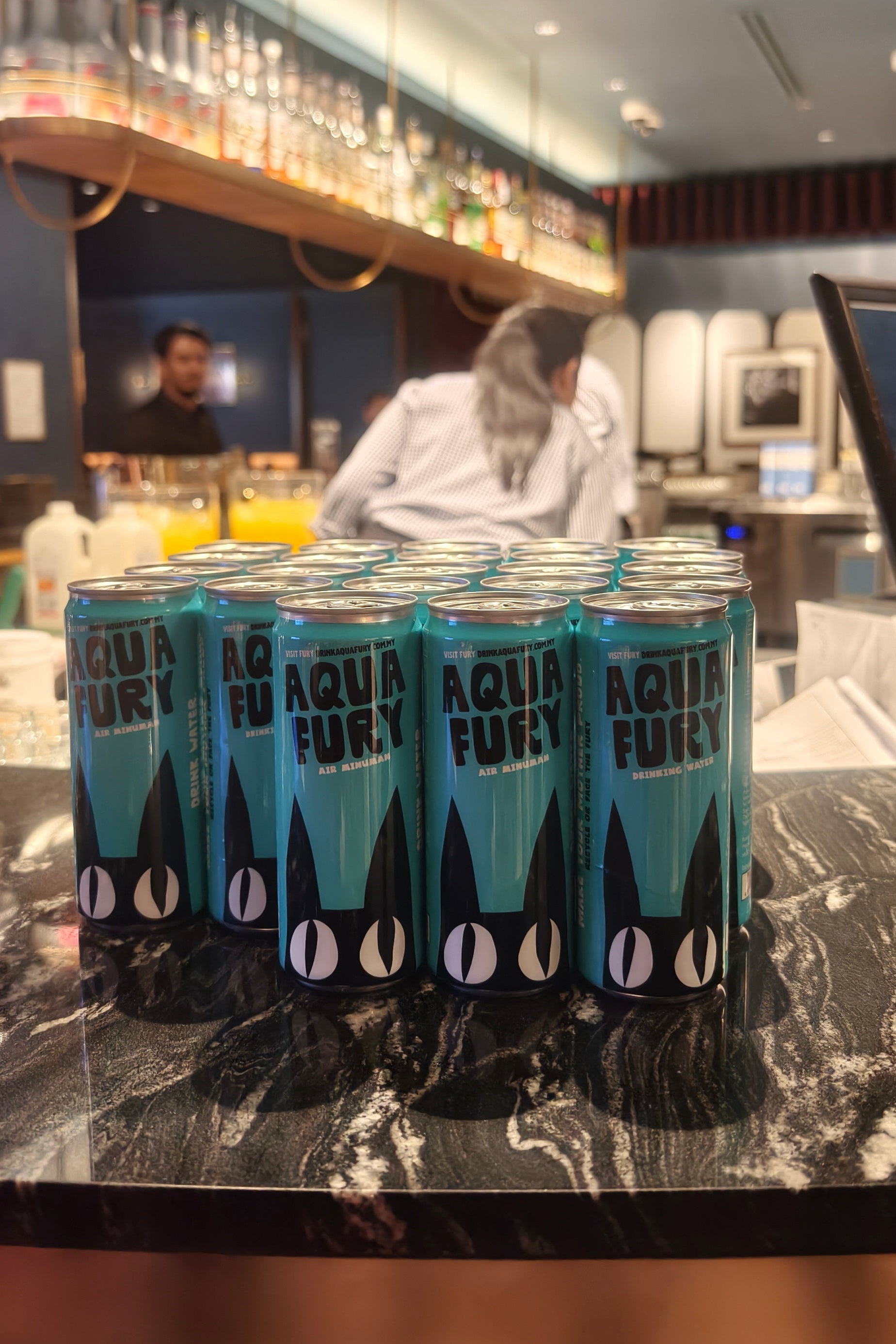 Aqua Fury cans on a bar counter with a blurred background of people and drinks.